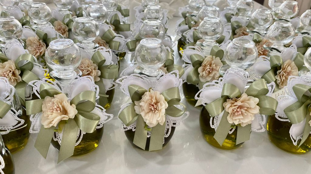 In questa foto si vede una delle Bomboniere di Sabrina composta da piccole bottiglie in vetro contenenti olio, decorate con fiocchi verde salvia, pizzo bianco e un fiore color crema applicato frontalmente.
