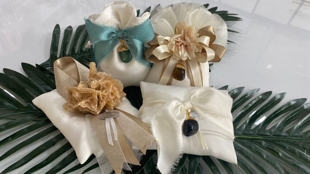 In questa foto si vede una delle Bomboniere di Sabrina realizzata con cuscinetti in tessuto chiaro, decorati con nastri in seta, fiori ornamentali e ciondoli in pietra naturale. Le bomboniere olistiche sono appoggiate su foglie decorative verdi.