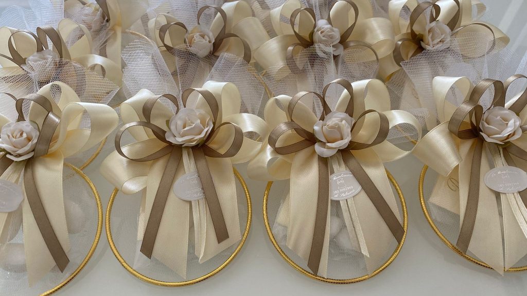 In questa foto si vede una delle Bomboniere di Sabrina composta da un tulle trasparente raccolto a sacchetto, decorato con un grande fiocco in nastro avorio e tortora e una rosa decorativa centrale. La bomboniera è rifinita con un anello dorato alla base e un cartellino personalizzato.