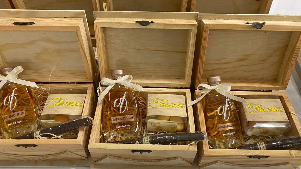 In questa foto si vede una delle Bomboniere di Sabrina presentata in una scatola di legno naturale con coperchio aperto. All’interno sono disposti una bottiglia di grappa, una scatola di confetti personalizzata e un sigaro, adagiati su paglia decorativa.