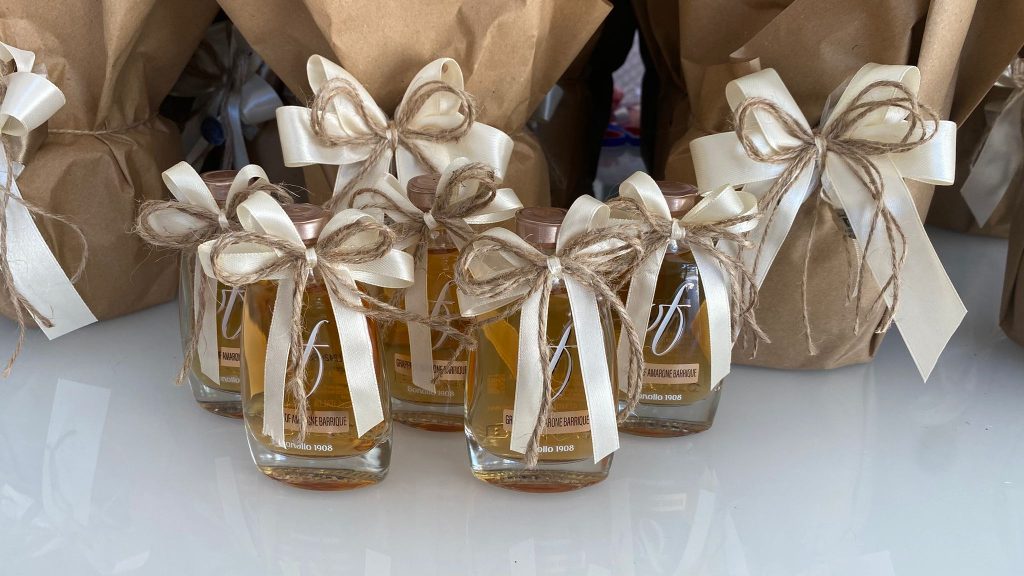 In questa foto si vede una delle Bomboniere di Sabrina con piccole bottiglie di liquore, decorate con fiocchi in nastro avorio e spago naturale. Le bottiglie sono disposte in fila su una superficie chiara, con sacchetti in carta sullo sfondo.