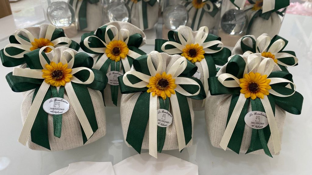 In questa foto si vede una delle Bomboniere di Sabrina realizzata con sacchetti in tessuto grezzo, chiusi da un fiocco verde scuro e avorio e decorati con un fiore giallo centrale. Ogni bomboniera presenta un cartellino con il logo.