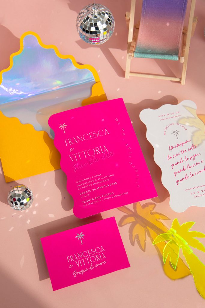 In questa foto si vede una delle tendenze partecipazioni matrimonio 2026: uno stile pop e irriverente, con cartoncini fucsia dalla forma sagomata, accompagnati da buste giallo fluo con interno olografico. I testi giocano con font moderni, citazioni e dettagli tropicali, come la palma e i mini specchi a forma di orsetto. L’allestimento include anche una mini sdraio arcobaleno e disco ball: un invito che trasmette gioia, leggerezza e spirito party.