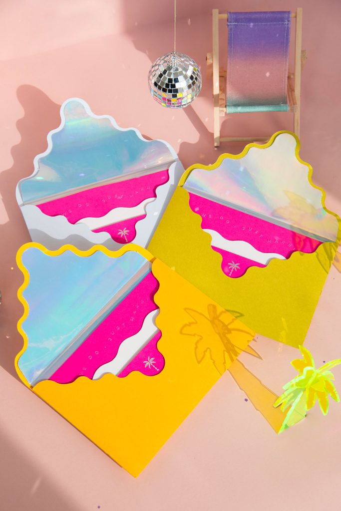 In questa foto si vede una delle tendenze partecipazioni matrimonio 2026: buste giallo limone e grigio perla con chiusura ondulata, che custodiscono inviti fucsia dai bordi sagomati. L’interno delle buste ha un rivestimento iridescente. L’insieme è completato da decorazioni tropicali come palme trasparenti e mini disco ball. Il tono è giocoso e moderno, perfetto per un matrimonio non convenzionale e colorato.