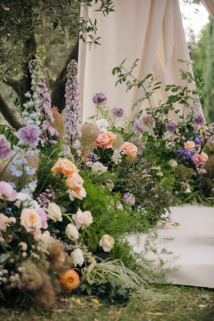 In questa foto si vede una delle tendenze Allestimenti floreali 2026: una scenografia nuziale immersa nel verde, con un tappeto di fiori che ricorda un giardino spontaneo. Tra le tonalità prevalgono il pesca, il lilla, il bianco e il rosa tenue. I fiori sono disposti ai lati di un cammino cerimoniale, incorniciato da drappi chiari e piante verdi, per un effetto naturale e incantato.