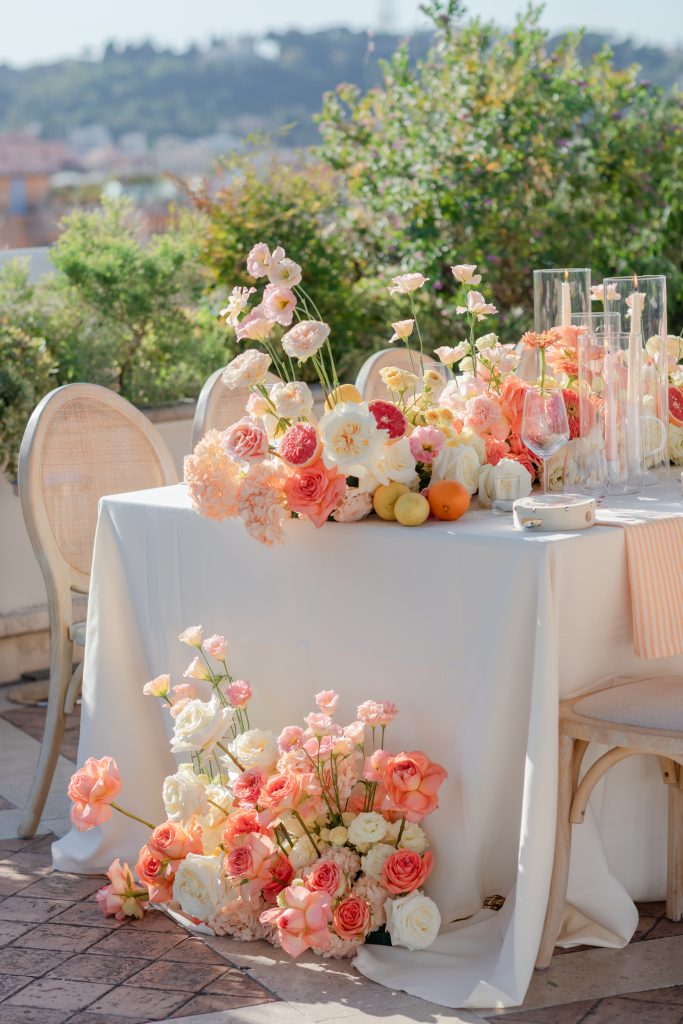 In questa foto si vede una delle tendenze Allestimenti floreali 2026: una tavola allestita all’aperto con composizioni floreali dai toni pesca, corallo, rosa e crema, posizionate sia sopra che alla base del tavolo. La scenografia è completata da frutta fresca sparsa tra i fiori, creando un effetto naturale e vibrante.