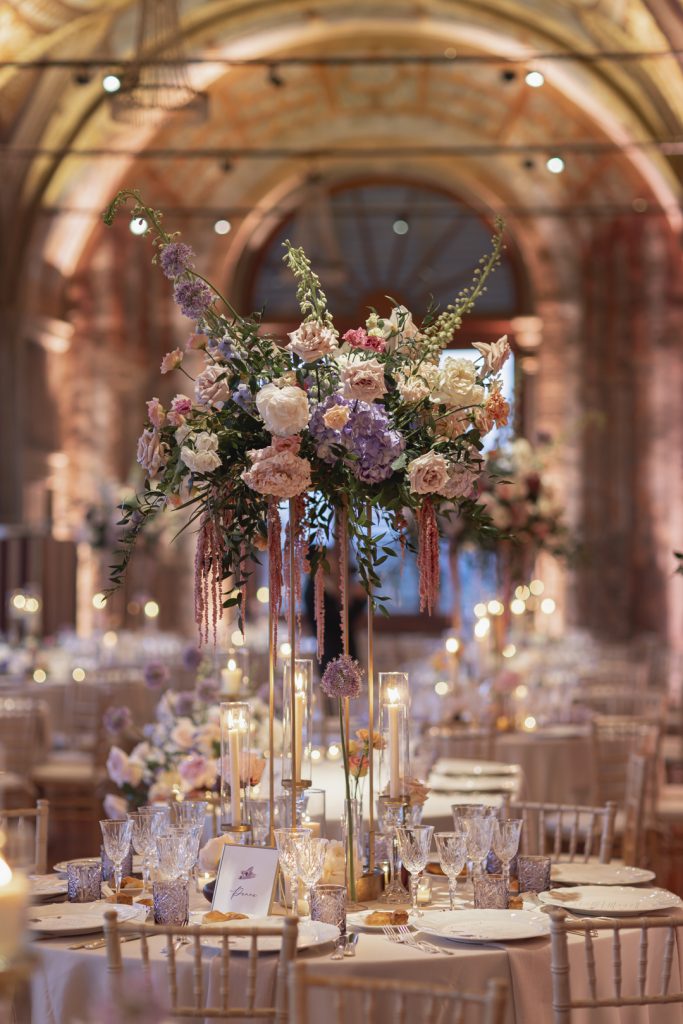 In questa foto si vede una delle tendenze Allestimenti floreali 2026: un centro tavola sospeso, scenografico e verticale, con fiori dai toni cipria, lilla, rosa e avorio, arricchito da elementi pendenti. L’atmosfera è resa ancora più elegante da una mise en place raffinata con calici in cristallo e candele alte.