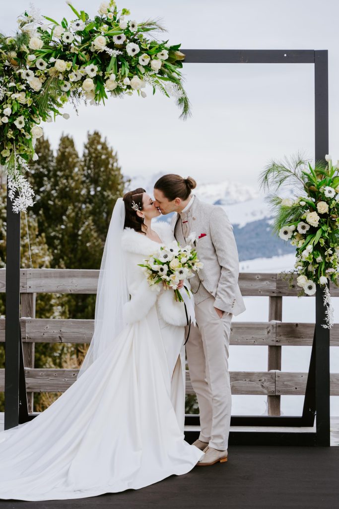 In questa foto si vede una delle tendenze Allestimenti floreali 2026: un arco moderno quadrato, decorato con fiori bianchi e verde brillante, incornicia il bacio degli sposi in una cerimonia invernale all’aperto. La sposa indossa un abito con maniche in pelliccia bianca, e lo sfondo mostra le montagne innevate, creando un contrasto tra gelo e romanticismo.