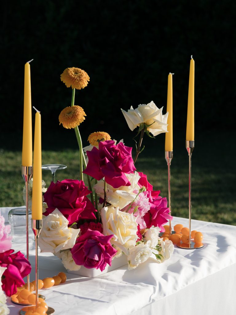 In questa foto si vede una delle tendenze Allestimenti floreali 2026: un centro tavola brillante e pop, composto da rose fucsia, rose bianche, e gerbere arancioni. Attorno, candele gialle alte e sfere decorative in tinta solare. Il contrasto tra colori accesi e sfondo verde prato crea un effetto grafico e contemporaneo.