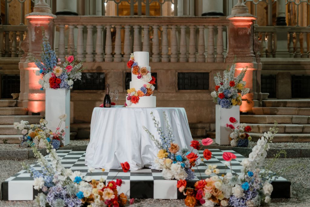 In questa foto si vede una delle tendenze Allestimenti floreali 2026: il tavolo della torta nuziale diventa protagonista scenico. Su una pedana a scacchi, la torta a piani bianchi è decorata con fiori colorati e vivaci, disposti anche intorno alla base. L’allestimento unisce elementi classici e pop, creando un impatto visivo audace e sofisticato.