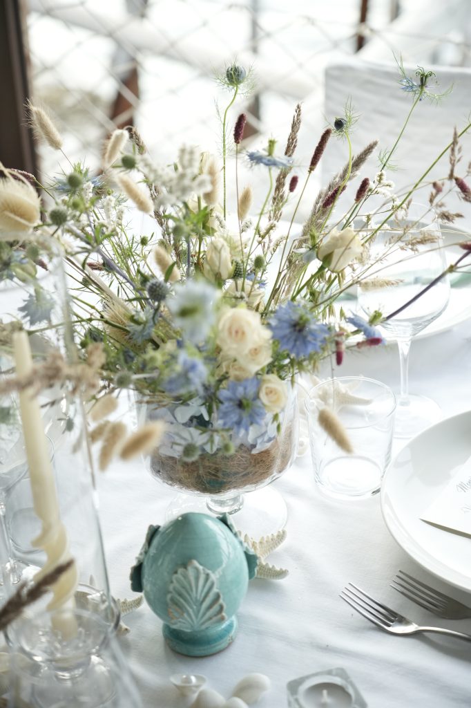 In questa foto si vede una delle tendenze Allestimenti floreali 2026: un centrotavola in stile wild e mediterraneo con fiori blu e panna, erbe essiccate e dettagli marini. Il centrotavola è racchiuso in una coppa trasparente, completato da elementi decorativi come una pigna in ceramica e conchiglie, per un mood naturale e costiero.