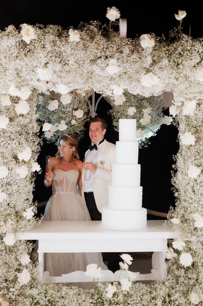 In questa foto si vede una delle tendenze Allestimenti floreali 2026: una scenografia notturna per il taglio torta, dove gli sposi sono circondati da un trionfo di gypsophila bianca (velo da sposa) e rose avorio. Il tavolo bianco è sormontato da una torta a più piani total white, enfatizzata da un allestimento floreale total white che dona un effetto nuvola.