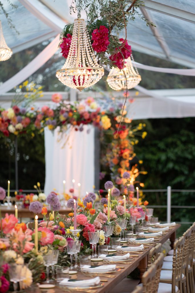 In questa foto si vede una delle tendenze Allestimenti floreali 2026: un ricevimento allestito sotto una tensostruttura trasparente, illuminata da lampadari con cristalli e fiori fucsia. Il lungo tavolo è decorato con un’esplosione di tulipani, peonie e fiori di campo nei toni del corallo, giallo e lilla, per un effetto vivace e festoso.