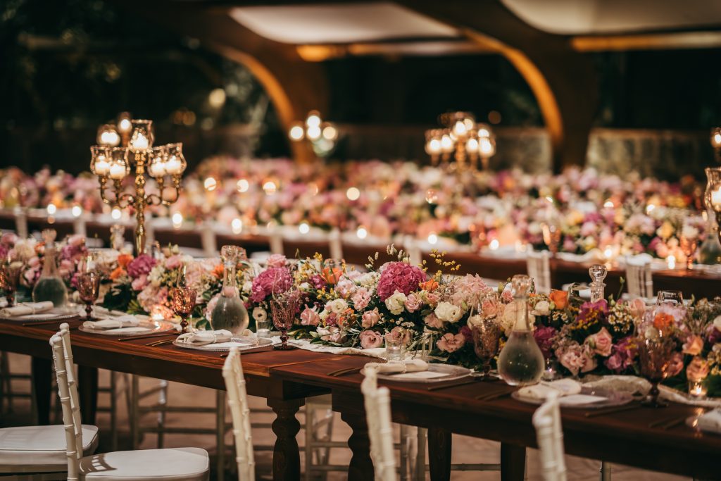 In questa foto si vede una delle tendenze Allestimenti floreali 2026: tavoli imperiali in legno scuro con allestimenti ricchi di fiori rosa, lilla e avorio. Le composizioni sono basse e fitte, intervallate da candelabri dorati e luci calde, creando un’atmosfera sofisticata e romantica.