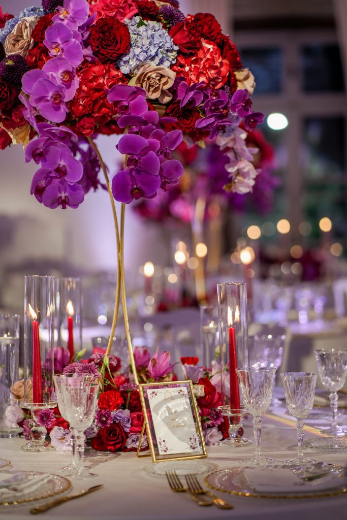 In questa foto si vede una delle tendenze Allestimenti floreali 2026: il ritorno del colore saturo con composizioni floreali ricche e intense, dominate da orchidee viola, rose rosse e dettagli color porpora, abbinate a candele rosse e calici in cristallo per un effetto luxury e teatrale.