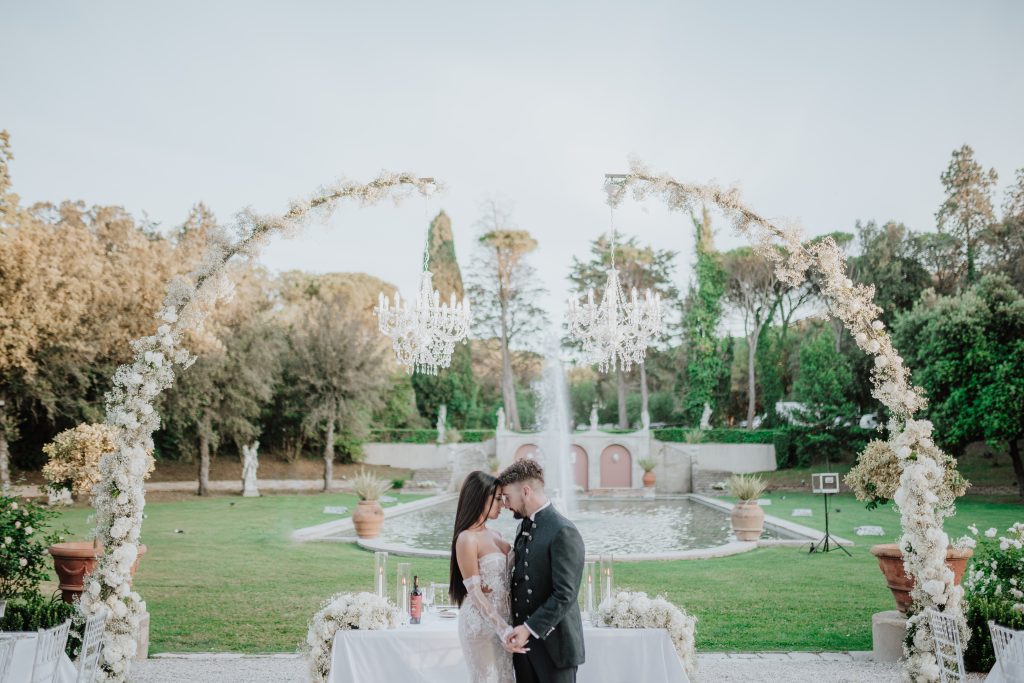 In questa foto si vede una delle tendenze Allestimenti floreali 2026: archi eterei di fiori bianchi a cornice della cerimonia all’aperto, impreziositi da lampadari pendenti che regalano un tocco romantico e sospeso nel tempo.