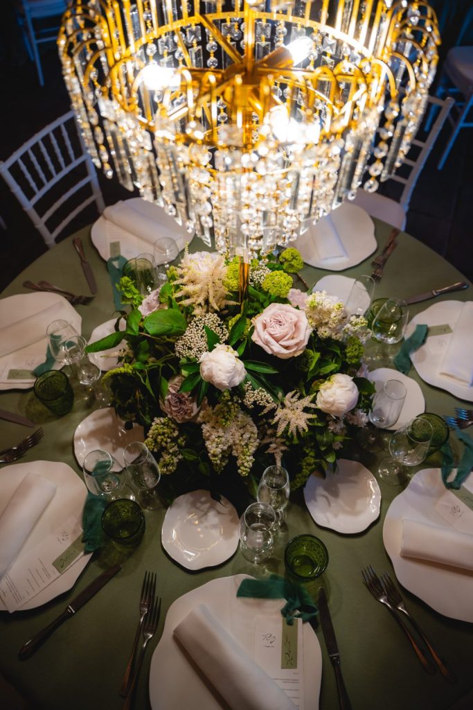 In questa foto si vede una delle tendenze Allestimenti floreali 2026: un centrotavola compatto e rigoglioso, con fiori bianchi, verdi e cipria, valorizzato da un grande lampadario dorato con pendenti in cristallo posizionato proprio sopra il tavolo. L’effetto è quello di un giardino lussureggiante illuminato da una luce preziosa.