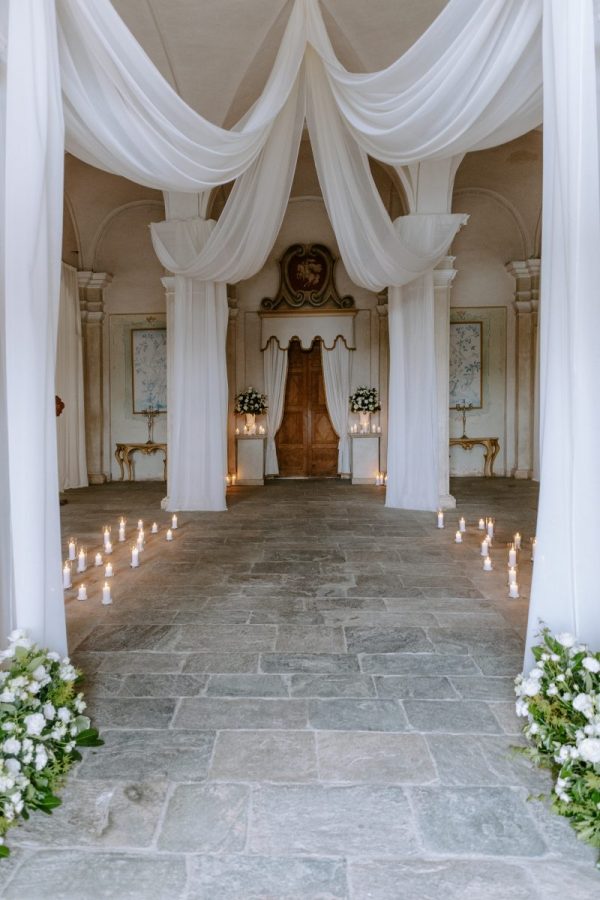 In questa foto si vede una delle tendenze allestimenti location matrimonio 2026: una sala interna allestita con drappi bianchi appesi al soffitto e candele disposte lungo il corridoio centrale. Ai lati, fiori bianchi e dettagli eleganti ricreano un’atmosfera sospesa e intima.