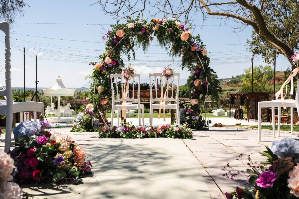 In questa foto si vede una delle Tendenze allestimenti matrimonio 2026: una lunga tavolata immersa nel verde, impreziosita da una spettacolare cascata di fiori freschi nelle tonalità del bianco, rosa cipria e malva, che dal centro del tavolo scivola fino al prato. Sedie in legno con seduta in paglia e veli bianchi appesi agli alberi creano un’atmosfera bucolica, romantica e leggera.