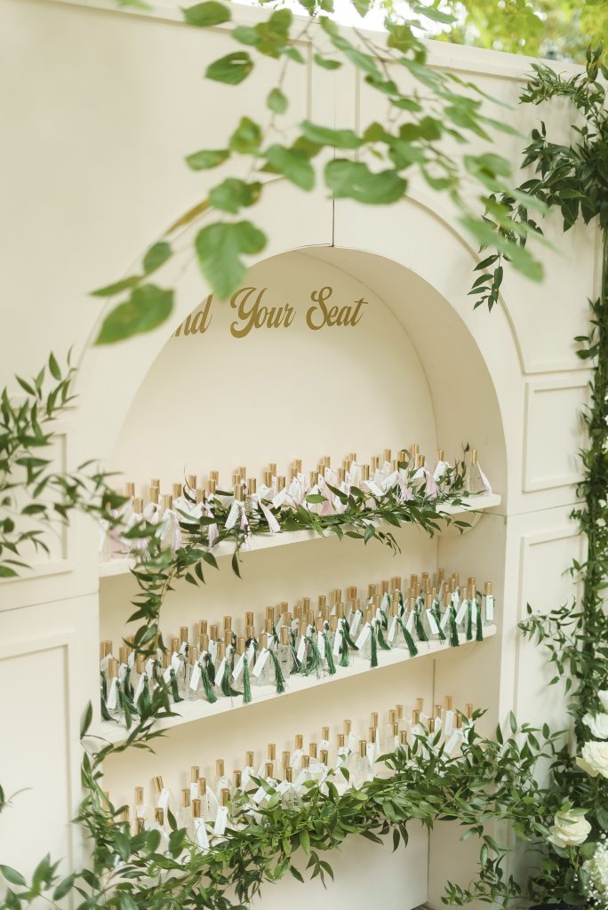 In questa foto si vede una delle tendenze allestimenti location matrimonio 2026: un tableau de mariage innovativo, realizzato con scaffali ad arco pieni di profumatori per ambienti personalizzati. Ogni bottiglietta ha una targhetta con il nome dell’ospite, inserita tra decorazioni di foglie verdi naturali.