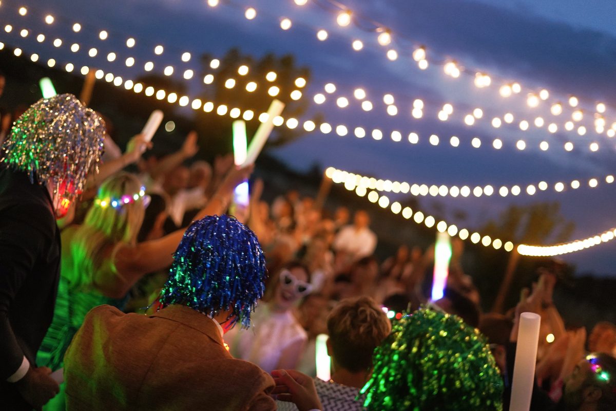 In questa foto si vede una delle tendenze allestimenti location matrimonio 2026: una festa serale all’aperto sotto un cielo di lucine sospese. Gli ospiti, in clima giocoso e festoso, indossano parrucche colorate e accessori luminosi, mentre ballano con bastoncini LED.