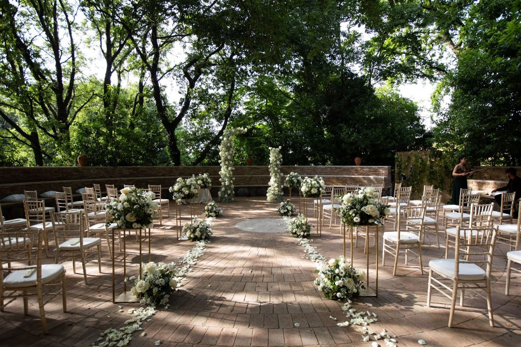 In questa foto si vede una delle tendenze allestimenti location matrimonio 2026: una cerimonia intima allestita in un contesto boschivo, con sedie chiare in legno e composizioni di fiori bianchi lungo il corridoio centrale. La natura fa da cornice a un allestimento elegante e semplice.