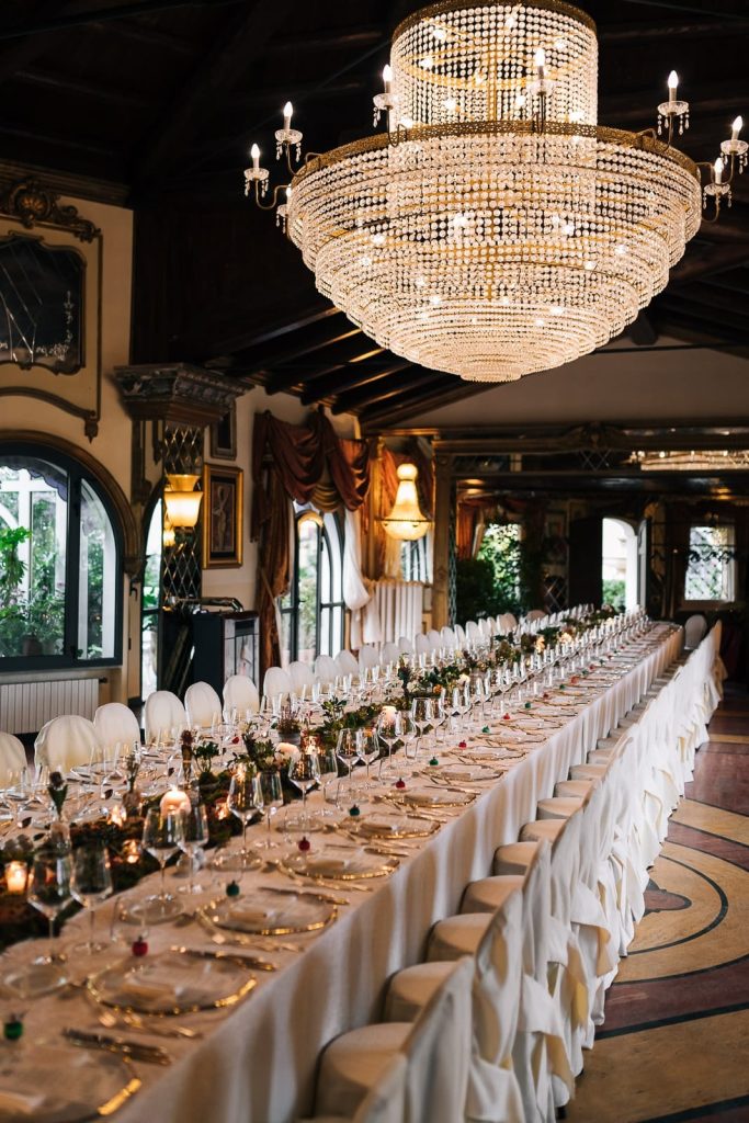 In questa foto si vede una delle tendenze allestimenti location matrimonio 2026: una lunga tavolata imperiale all’interno di una sala dallo stile classico, con un grande lampadario di cristallo, mise en place raffinata e decorazioni verdi e dorate che percorrono il centro tavola.