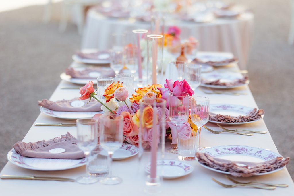 In questa foto si vede una delle tendenze allestimenti location matrimonio 2026: una tavola elegante con piatti decorati, tovaglioli rosa cipria, calici trasparenti e un centrotavola floreale nelle sfumature del rosa, lilla e arancio. La palette è calda, romantica e contemporanea.