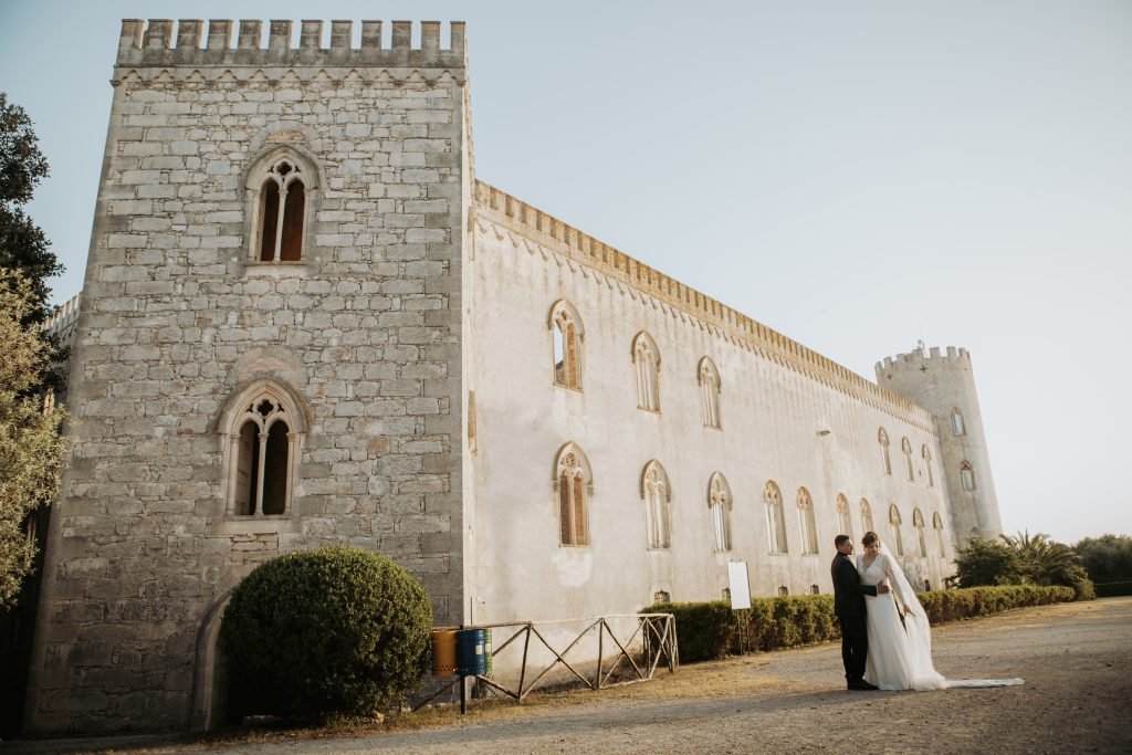 In questa foto si vede una delle fotografie di matrimonio di Davide Meli: una coppia di sposi è ritratta all’esterno di un edificio storico in pietra, con torri e finestre ad arco; i due si tengono per mano in uno spazio ampio e silenzioso, immerso nella luce del tardo pomeriggio.