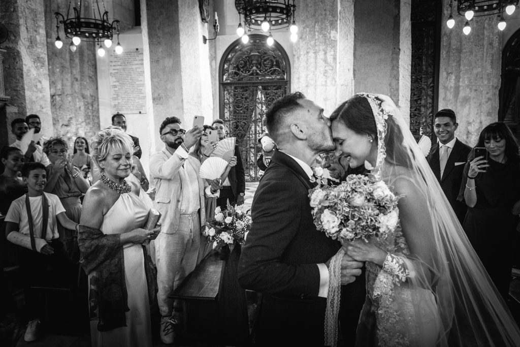 In questa foto si vede una delle fotografie di matrimonio di Davide: gli sposi si baciano durante la cerimonia in chiesa, circondati dagli invitati; l’immagine in bianco e nero cattura l’emozione del momento e le reazioni partecipi delle persone intorno.