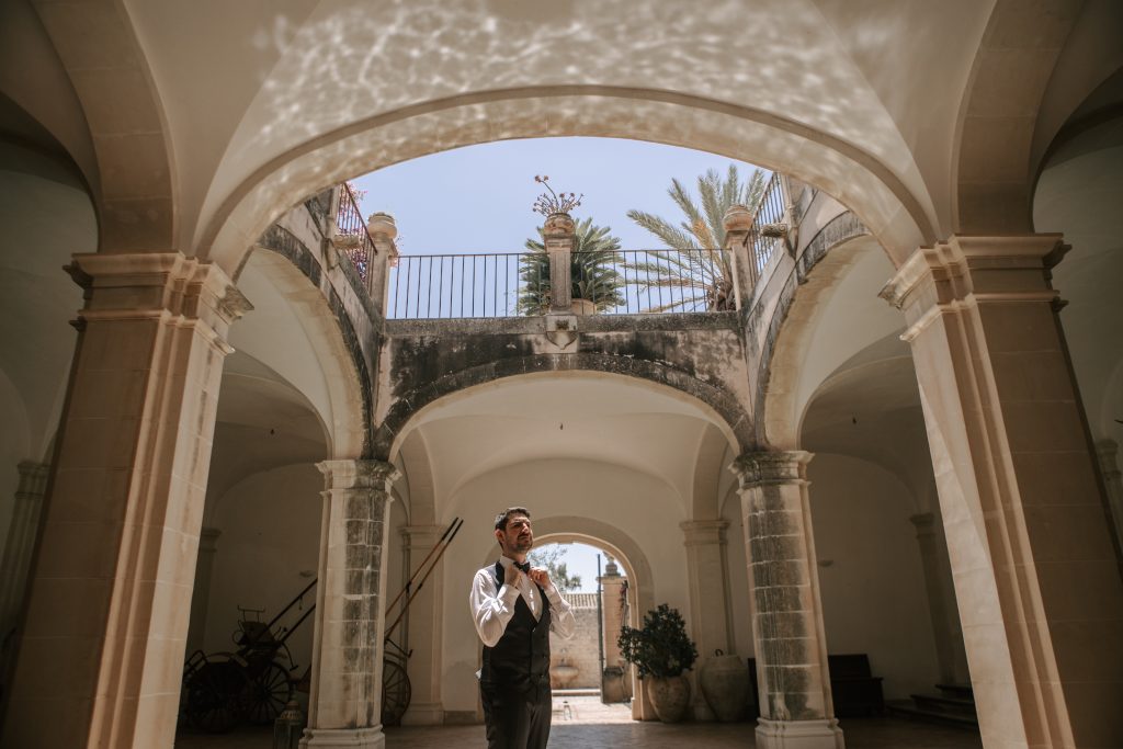 In questa foto si vede una delle fotografie di matrimonio di Davide Meli: lo sposo è solo in un cortile storico con archi e colonne in pietra, mentre sistema il papillon; la scena è illuminata dalla luce naturale che entra dall’alto, creando un’atmosfera calma e raccolta.