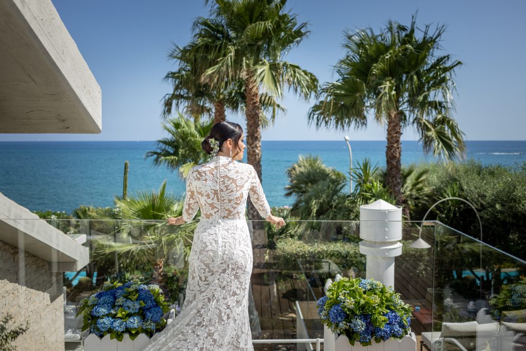 In questa foto si vede una delle fotografie di matrimonio del fotografo Meli, una sposa ripresa di spalle su una terrazza affacciata sul mare, con abito in pizzo a maniche lunghe, palme e acqua azzurra sullo sfondo.