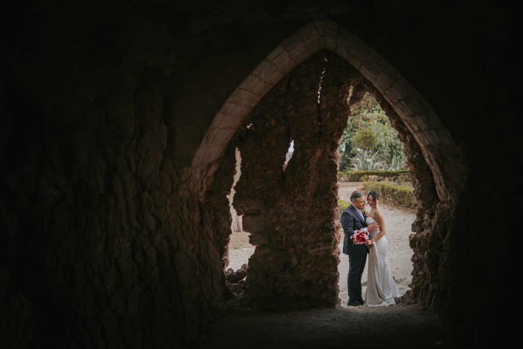 In questa foto si vede una delle fotografie di matrimonio di Davide Meli: gli sposi sono abbracciati all’ingresso di un antico arco in pietra, osservati dall’interno di una struttura buia che incornicia la scena e crea un forte contrasto tra ombra e luce naturale.