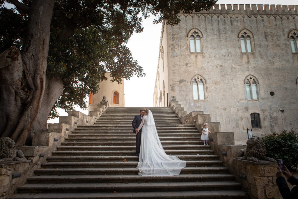 In questa foto si vede una delle fotografie di matrimonio di Davide Meli, due sposi fermi al centro di una grande scalinata in pietra davanti a un edificio storico, con una bambina visibile sul lato destro dei gradini.