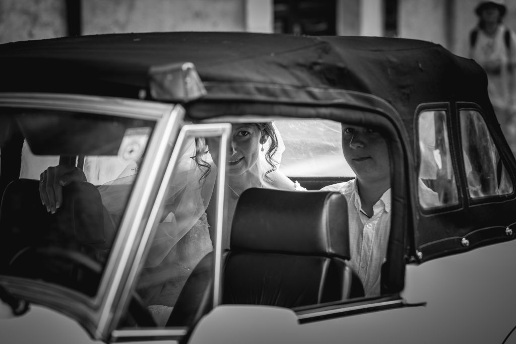 In questa foto si vede una delle fotografie di matrimonio di Davide Meli: la sposa è seduta all’interno di un’auto d’epoca scoperta, in bianco e nero, mentre guarda fuori dal finestrino; accanto a lei un’altra persona sul sedile posteriore, in un momento intimo e sospeso prima della cerimonia.