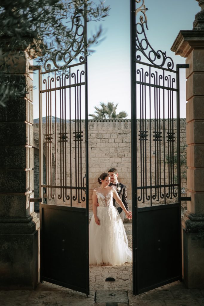 In questa foto si vede una delle fotografie di matrimonio di Davide Meli, una coppia di sposi ripresa attraverso un grande cancello in ferro aperto, con muri in pietra chiara e cielo azzurro sullo sfondo.