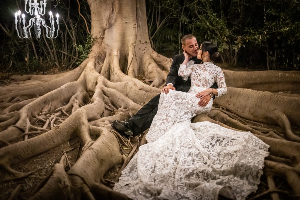 In questa foto si vede una delle fotografie di matrimonio di Davide Meli: gli sposi sono seduti sulle grandi radici di un albero monumentale, illuminati da lampadari appesi tra i rami; si guardano da vicino in un’atmosfera notturna, naturale e suggestiva.