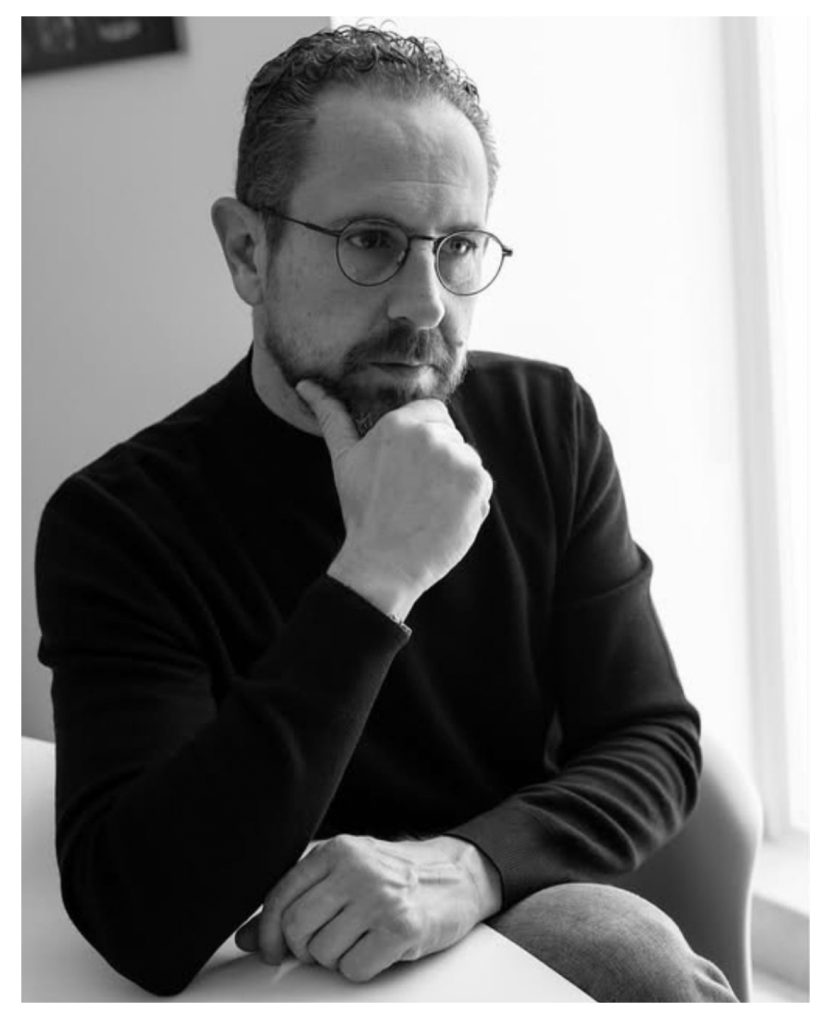 In questa foto si vede una fotografia di Davide Meli, un ritratto in bianco e nero dell' uomo seduto, con occhiali, barba corta e mano appoggiata al mento, illuminato lateralmente da una finestra.