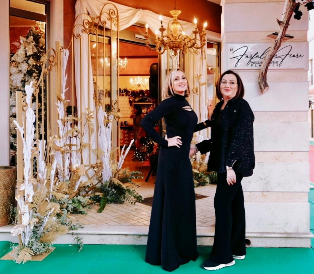 In questa foto si vede uno degli allestimenti de Le Farfalle in Fiore - Angela Crupi: due donne in total black sono ritratte all’ingresso di uno showroom elegante, con una scenografia dorata, dettagli in bianco e verde e decorazioni natalizie sullo sfondo. L’insegna dell’atelier è ben visibile sulla destra.