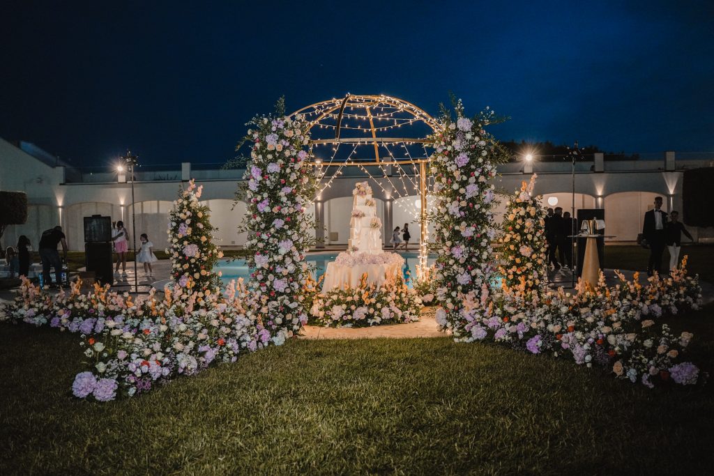 In questa foto si vede uno degli allestimenti de Le Farfalle in Fiore - Angela Crupi: in un giardino notturno illuminato da fili di luci, una wedding cake è incorniciata da fiori pastello disposti a semicerchio. Il prato e la piscina fanno da sfondo all’elegante struttura a cupola.