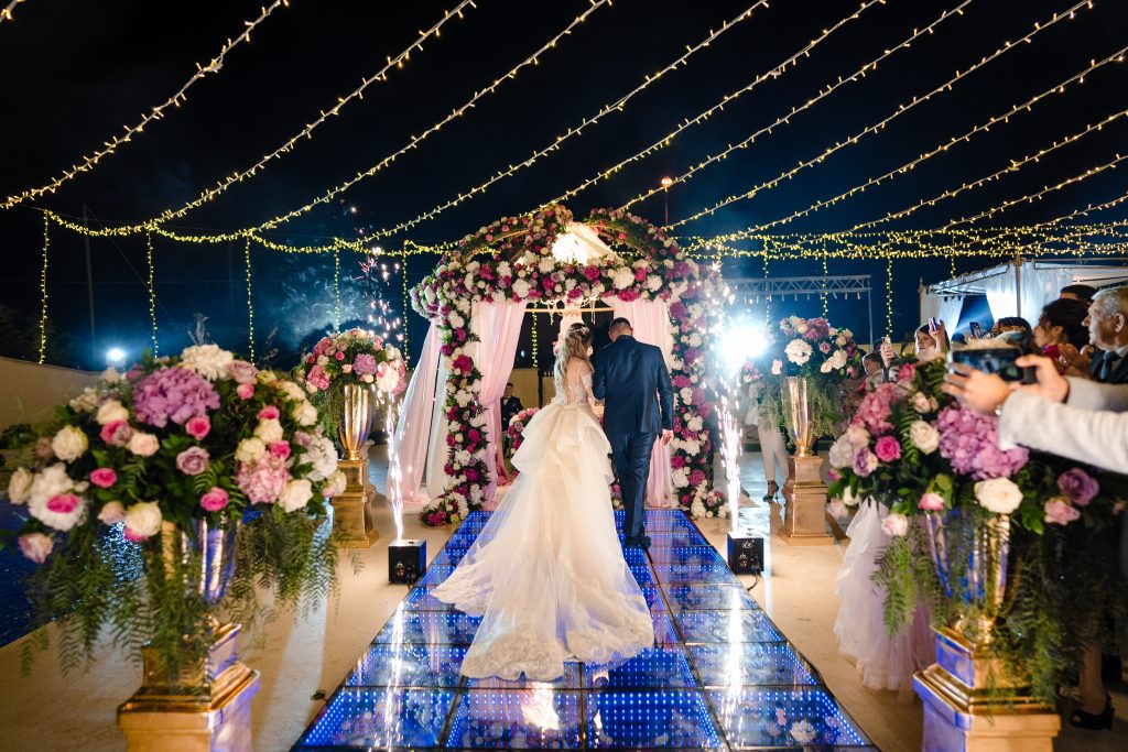 In questa foto si vede uno degli allestimenti de Le Farfalle in Fiore - Angela Crupi: gli sposi percorrono una passerella luminosa, circondata da colonne dorate e vasi stracolmi di ortensie e rose in toni rosa e bianchi. Sotto un cielo stellato di luci a filo e fuochi d’artificio, entrano in un gazebo ricoperto di fiori, per un effetto scenografico altamente emozionale.