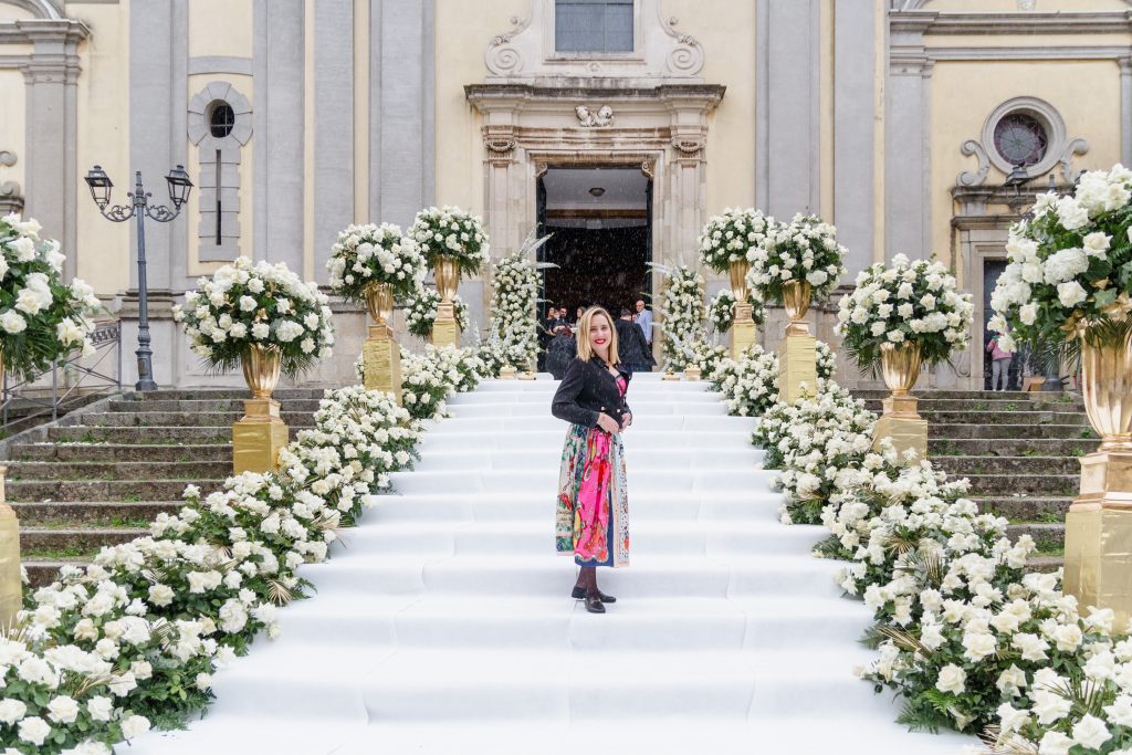 In questa foto si vede uno degli allestimenti de Le Farfalle in Fiore - Angela Crupi: una scalinata monumentale ricoperta di moquette bianca è fiancheggiata da decine di composizioni floreali total white con rose e ortensie, inserite in eleganti vasi dorati. Sullo sfondo, la facciata barocca di una chiesa e la wedding planner in posa sorridente.