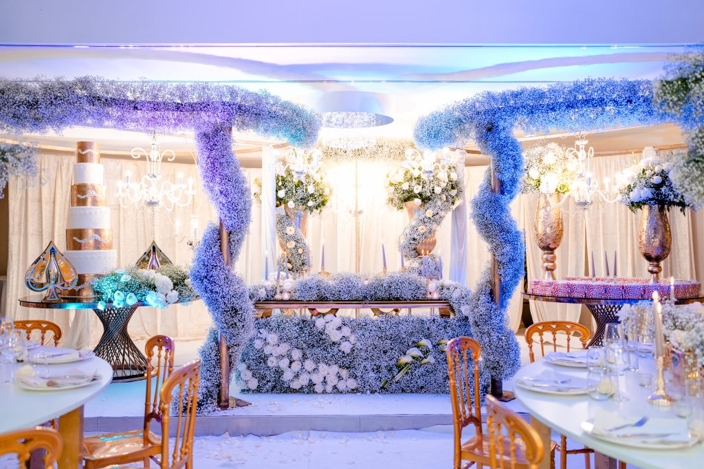 In questa foto si vede uno degli allestimenti de Le Farfalle in Fiore - Angela Crupi: l’interno di una sala ricevimenti si trasforma in un regno incantato con strutture ricoperte interamente da nuvole di gypsophila azzurra, candide rose, chandelier e dettagli dorati. L’effetto è futuristico ma fiabesco, con una mise en place essenziale su tavoli tondi.