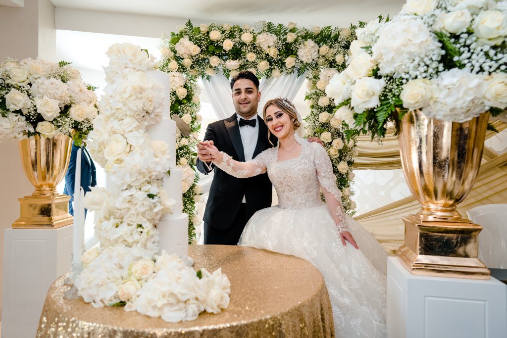 In questa foto si vede uno degli allestimenti de Le Farfalle in Fiore – Angela Crupi
Una coppia di sposi sta tagliando una torta nuziale bianca a più piani, decorata con fiori chiari e scenografici. La sposa indossa un abito principesco in pizzo con maniche lunghe e un'acconciatura raccolta con dettagli gioiello; lo sposo è in smoking nero con papillon. Alle loro spalle un grande arco fiorito bianco e verde crea una cornice romantica, mentre due grandi vasi dorati con composizioni floreali arricchiscono la scena. Il tavolo rotondo, coperto da una tovaglia oro glitterata, completa un’atmosfera elegante e regale.