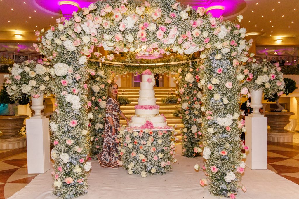 In questa foto si vede uno degli allestimenti de Le Farfalle in Fiore - Angela Crupi: una maxi torta a cinque piani è incorniciata da un tunnel floreale total white e rosa in gypsophila e rose, per un effetto scenografico sontuoso e romantico. Al centro, Angela Crupi sorride accanto alla sua creazione.