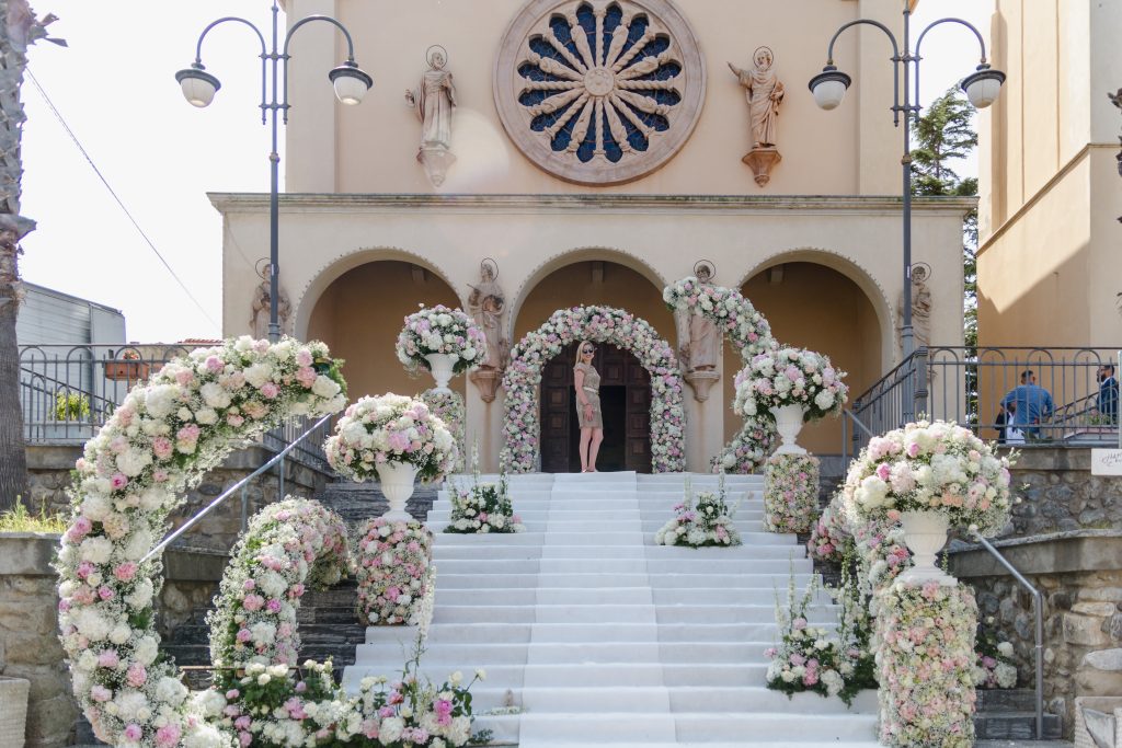 In questa foto si vede uno degli allestimenti de Le Farfalle in Fiore - Angela Crupi: la facciata di una chiesa con tre arcate è incorniciata da composizioni floreali a spirale in rosa e bianco, disposte su colonne e a terra. La wedding planner posa al centro della scalinata bianca, tra gli archi fioriti.