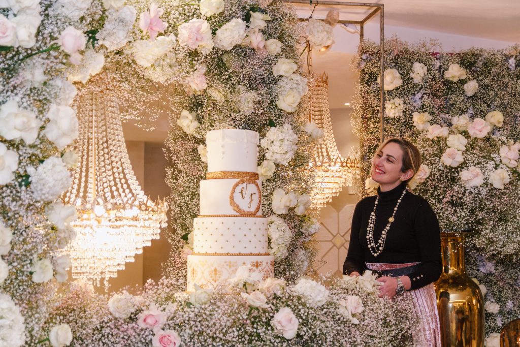 In questa foto si vede uno degli allestimenti de Le Farfalle in Fiore – Angela Crupi
La wedding designer osserva sorridente una torta nuziale a tre piani bianca e oro, posizionata al centro di una scenografia floreale ricchissima. L’ambiente è decorato con una profusione di fiori bianchi e rosa cipria, tra nuvole di gypsophila e ortensie. Sullo sfondo, due grandi lampadari a goccia in cristallo illuminano lo spazio con riflessi lussuosi. L’atmosfera è fiabesca, sofisticata e romantica.