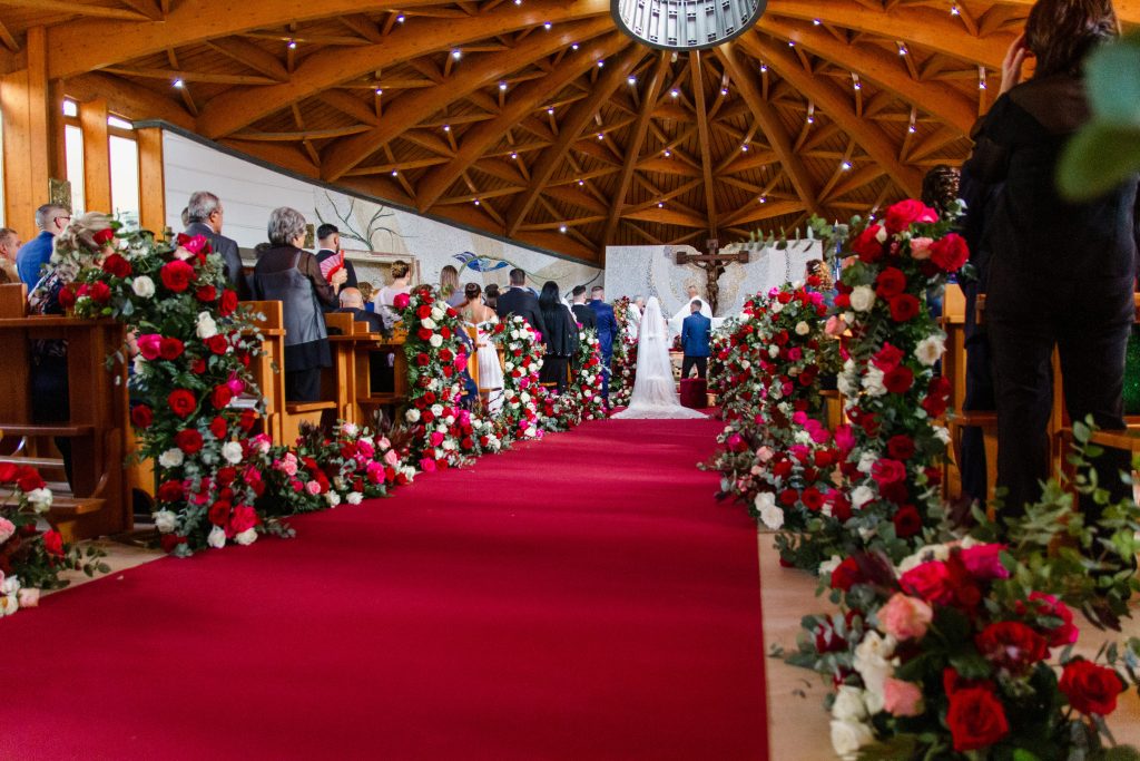 In questa foto si vede uno degli allestimenti de Le Farfalle in Fiore - Angela Crupi: all’interno di una chiesa moderna, una lunga navata rossa è affiancata da composizioni abbondanti di rose rosse e bianche. Gli sposi si avviano all’altare tra l’affetto degli invitati, per una cerimonia dall’impatto visivo molto forte.