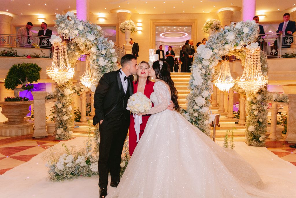 In questa foto si vede uno degli allestimenti de Le Farfalle in Fiore - Angela Crupi: gli sposi baciano la wedding planner all’interno di un’area ricevimenti decorata con archi fioriti, chandelier e dettagli luxury. L’atmosfera è elegante, calda e gioiosa, tra luci soffuse e dettagli curati in ogni angolo.