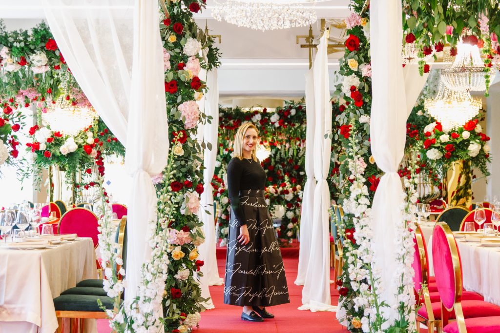 In questa foto si vede uno degli allestimenti de Le Farfalle in Fiore - Angela Crupi: un corridoio scenografico interno decorato con colonne floreali bianche, rosse e rosa, tende di voile e sedute color magenta. Al centro, la wedding planner posa sorridente con un outfit nero.